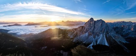 Foto de Salida del sol sobre el pico de la montaña Sass de Puta en Passo delle Erbe pasar contra los picos de Dolomita en el fondo, cubierta de nubes inversa en el valle, los rayos del sol. Panorama de paisaje montañoso de drones aéreos. - Imagen libre de derechos