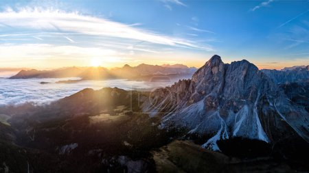 Foto de Salida del sol sobre el pico de la montaña Sass de Puta en Passo delle Erbe pasar contra los picos de Dolomita en el fondo, cubierta de nubes inversa en el valle, los rayos del sol. Panorama de paisaje montañoso de drones aéreos. - Imagen libre de derechos