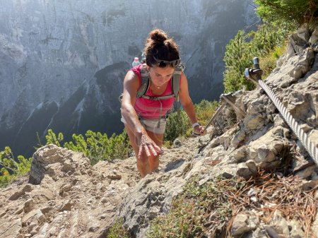 Téléchargez les photos : Femme sportive en trek dans les Dolomites, t-shirt rouge, short, cheveux foncés, sac à dos. Escalade physiquement exigeante, montagnes et forêts en arrière-plan. - en image libre de droit