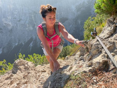 Téléchargez les photos : Femme plus âgée sportive en trek dans les Dolomites, t-shirt rouge, short, cheveux foncés, sac à dos. Escalade physiquement exigeante, montagnes et forêts en arrière-plan. - en image libre de droit
