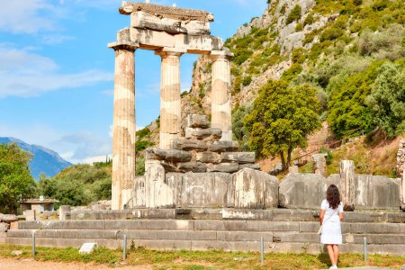 Eine langhaarige Frau im weißen Kleid blickt auf die antike Tempelanlage der Athena Pronaia in Delphi. Sonniger Tag, blauer Himmel. Die archäologische Stätte, UNESCO-Weltkulturerbe, Delphi, Griechenland.