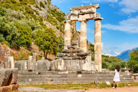 Eine langhaarige Frau im weißen Kleid blickt auf die antike Tempelanlage der Athena Pronaia in Delphi. Sonniger Tag, blauer Himmel. Die archäologische Stätte, UNESCO-Weltkulturerbe, Delphi, Griechenland.