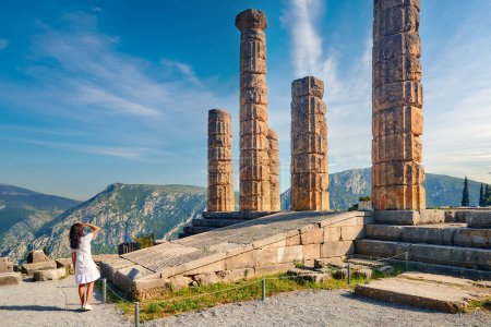 Eine langhaarige Frau blickt von hinten auf den Apollo-Tempel oder Apollonion und seine dorischen Säulen im Sonnenuntergang. Touristenort, berühmt für Orakel im Apollo-Heiligtum. Mount Parnassus, Delphi, Griechenland.