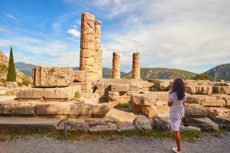 Eine langhaarige Frau blickt von hinten auf den Apollo-Tempel oder Apollonion und seine dorischen Säulen im Sonnenuntergang. Touristenort, berühmt für Orakel im Apollo-Heiligtum. Mount Parnassus, Delphi, Griechenland.