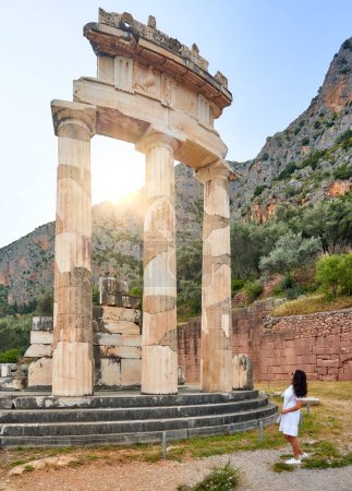 Eine langhaarige Frau im weißen Kleid blickt auf die riesigen Säulen der antiken Tempelanlage Athena Pronaia in Delphi. Sonniger Tag, blauer Himmel. Vertikales Foto, archäologisch. UNESCO Stätte, Delphi, Griechenland.