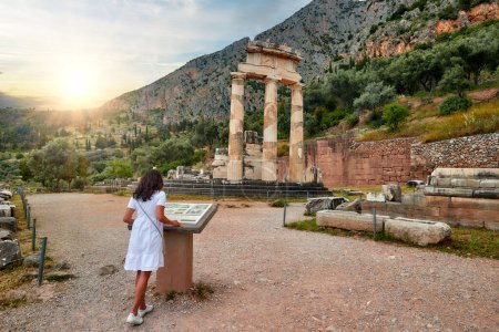 Eine langhaarige Frau in weißem Kleid liest Informationen von einer Tafel im antiken Tempelkomplex Athena Pronaia in Delphi. Sonnenuntergang, blauer Himmel. Archäologische Stätte, UNESCO-Weltkulturerbe.