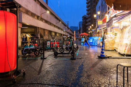 Foto de Osaka, Japón - 29 de abril de 2023: Scooters eléctricos y bicicletas estacionados en la brillante farmacia y estación de tren en la noche lluviosa. Foto de alta calidad - Imagen libre de derechos