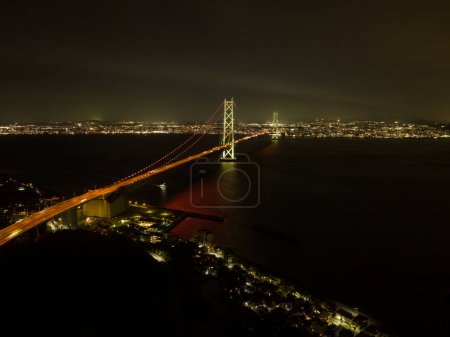 Photo for Akashi Kaikyo suspension bridge over dark water between city lights at night. High quality photo - Royalty Free Image