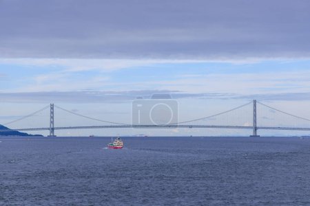 Photo for Small vessel sails calm blue waters by suspension bridge on cloudy day. High quality photo - Royalty Free Image