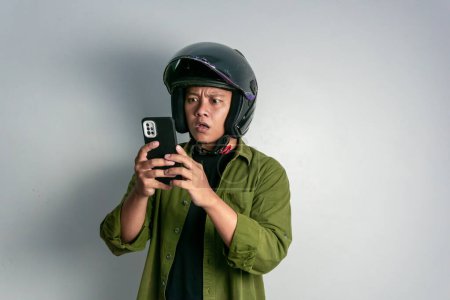 Foto de Hombre asiático adulto confuso al mirar a su teléfono mientras usa casco de motocicleta. Hombre mirando a su teléfono con expresión conmocionada - Imagen libre de derechos