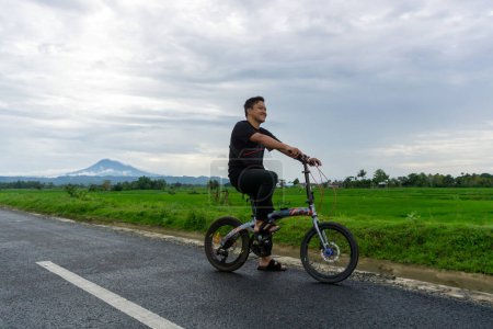 Feliz hombre asiático montando una bicicleta por la mañana en el camino de asfalto. Ciclismo con vista a la montaña y arrozal al fondo. 