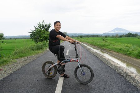 Feliz hombre asiático montando una bicicleta por la mañana en el camino de asfalto. Ciclismo con vista a la montaña y arrozal al fondo. 