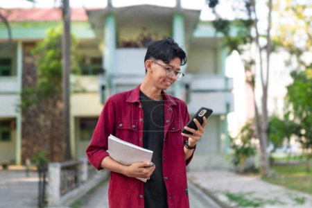 Photo for Young Asian college student using smartphone with happy expression. A male smiling while holding his phone and books at the public park. copy space - Royalty Free Image