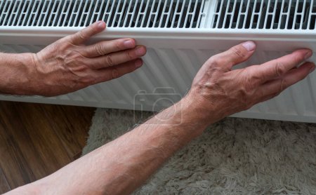 Foto de Primer plano de las manos de un hombre calentándose por el radiador de calefacción en el apartamento. El concepto de calefacción en la habitación. Una persona se calienta en casa tocando un radiador caliente con las manos. El hombre está congelado en casa.. - Imagen libre de derechos