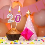 Decorations with balloons and a happy birthday candle with the number 20. Happy birthday greetings in pink flowers for a twenty-year-old girl, copy space. Muffin with a burning candle