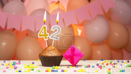 Téléchargez les photos : Decorations with balloons and a happy birthday candle with the number 42 for a woman. Happy birthday greetings in pink flowers for forty-two years old for an adult girl, copy space. Muffin with a burning candle - en image libre de droit