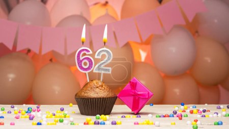 Téléchargez les photos : Decorations with balloons and a happy birthday candle with the number 62 for a woman. Happy birthday greetings in pink flowers for sixty-two years old for an adult woman, copy space. Muffin with a burning candle - en image libre de droit