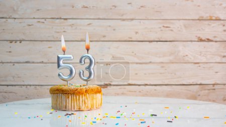 Téléchargez les photos : Happy birthday greetings for 53 years of silver numbers on the background of white boards copy space. Beautiful birthday card with a muffin with a burning candle for fifty-three years - en image libre de droit