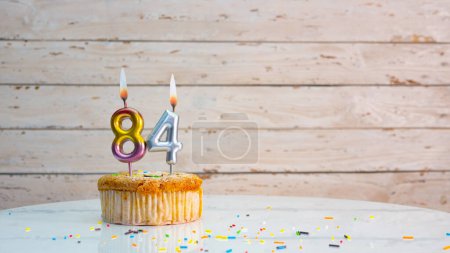 Photo for Happy 84th birthday greetings from silver numbers on white boards background copy space. Beautiful birthday card with a cupcake with a lit candle for eighty-four years. - Royalty Free Image