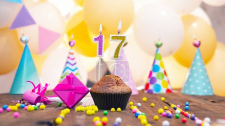 Photo for Happy birthday with a number of candles for seventeen years old against the background of balloons. A festive muffin with burning candles and a pink gift box for a girl. Happy birthday for 17 years old girl. - Royalty Free Image