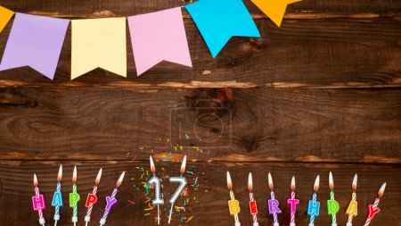 Photo for Top view, birthday decorations from letters of candles with fire and festive garlands, save space. Happy birthday background with number 17 on brown board table. Happy birthday to a seventeen year old - Royalty Free Image