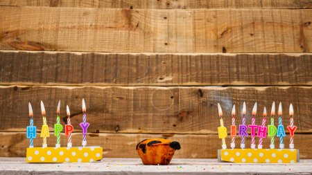 Foto de Muffin with a burning candle background for the anniversary on a beautiful wooden background. Postcard with letters of candles with congratulations for the holiday before the birthday. . Copy space. Happy birthday background - Imagen libre de derechos