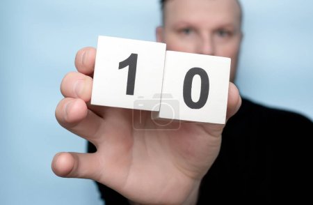 A young man in his hand holding a cube with the number 10 on a blue copy space background.
