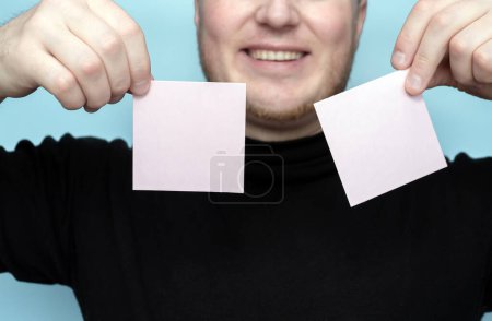 Foto de With a smile A young man holds two blank stickers for the inscription of your text. Message on sticker copy space. Mini banner for text in a person's hand. On a light pastel background - Imagen libre de derechos