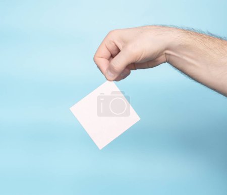 Foto de A hand holds a blank letter sticker for your text inscription. Message on sticker copy space. Mini banner for text in a person's hand. On a pastel blue background - Imagen libre de derechos