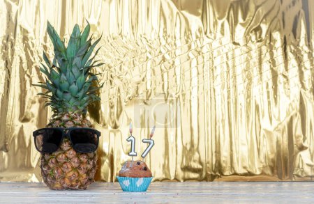 Photo for Cheerful pineapple in glasses festive happy birthday character with number  17. Beautiful background for congratulations Koiya space on a golden shiny background. Anniversary congratulations with the number of candles burning with a muffin. - Royalty Free Image