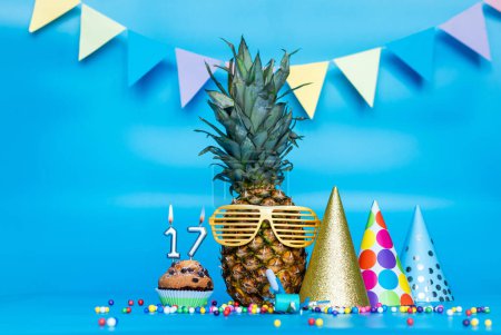 Photo for Creative background with pineapple character in sunglasses copy space. Happy birthday background with muffin or cake with candle number  17. Anniversary holiday decorations on a blue background. - Royalty Free Image