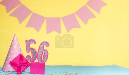 Geburtsdatum für ein Mädchen 56. Kopierraum. Geburtstag in Rosatönen vor gelbem Hintergrund. Dekorationen mit nummerierten Kerzen und einer Geschenkbox. Geburtstagskarte für eine Frau