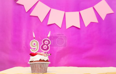 Background date of birth with number  98. Pink background with a cake and burning candles, save space, happy birthday anniversary for a girl. Holiday pudding muffin.