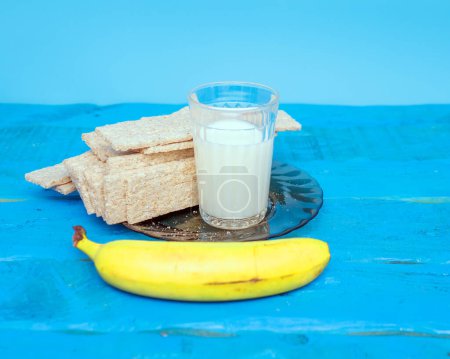 Pain sec avec lait et banane pour le petit déjeuner du matin. Un verre de lait avec une banane aux craquelins, sur une table de cuisine en bois.