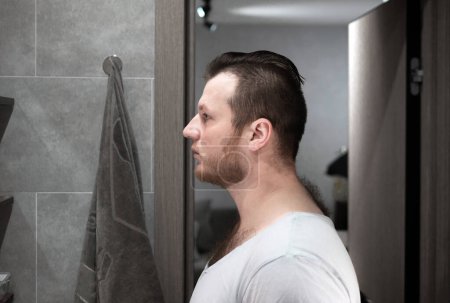 Sad man with a beard in the bathroom. Man looks in the mirror in the bathroom. Sad man at home.