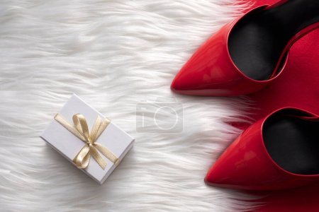 Photo for Close up top view of red ponty toe glossy shoes on white fuzzy faux fur, and a small white gift box with golden ribbon bowtie - Royalty Free Image