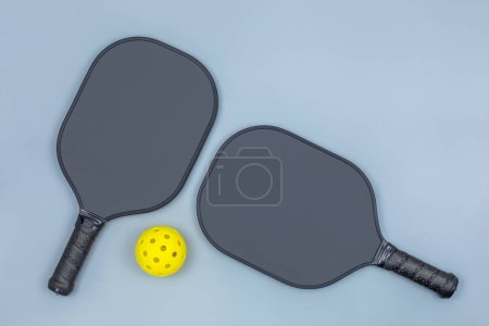Closeup top view of two black pickleball paddles and a yellow ball on light gray background with copy space