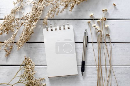Foto de Flatlay primer plano con un bloc de notas blanco blanco, pluma 0n pintado fondo de madera blanca con flores secas y hierbas que lo enmarcan. - Imagen libre de derechos