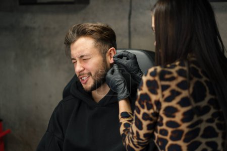 Téléchargez les photos : Studio de perçage de tatouage - femme fait un perçage d'oreille à un homme à un gars - en image libre de droit