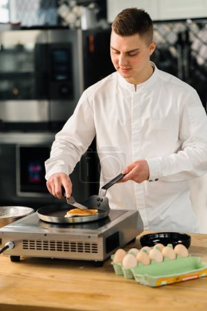 Foto de Un chef profesional en la cocina de un restaurante prepara panqueques para el desayuno. Receta culinaria Sartén en estufa - Imagen libre de derechos