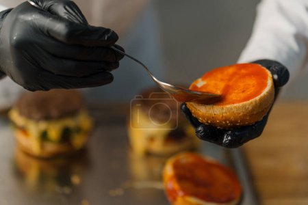 Téléchargez les photos : Un chef prépare un burger Gros plan de la main d'un homme étalant de la sauce rouge sur un pain frit - en image libre de droit