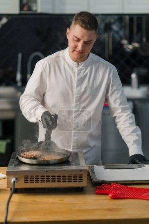 Téléchargez les photos : Chef en gants noirs prépare des galettes de hamburger savoureux poêle chaude dans la cuisine professionnelle - en image libre de droit
