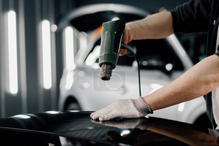 Téléchargez les photos : Un travailleur couvre un spoiler de voiture avec un film dans l'atelier à l'aide d'un sèche-cheveux et d'une spatule close-up Détails et soins de la voiture - en image libre de droit