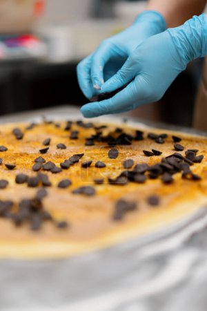 Foto de La cocina en la panadería - el proceso de fabricación de cinabriones, mujer panadero utiliza chocolate en gránulos para hornear - Imagen libre de derechos