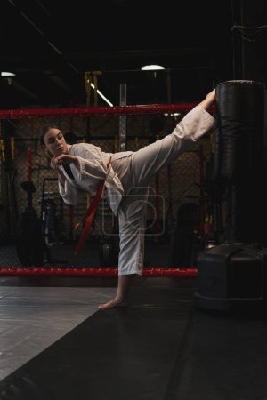 Téléchargez les photos : Une fille dans un kimono exerce avec un sac de boxe dans la salle de gym tout en apprenant les arts martiaux de karaté - en image libre de droit