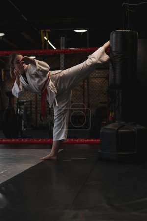 Foto de Una chica en un kimono hace ejercicios con un saco de boxeo en el gimnasio mientras aprende artes marciales de karate - Imagen libre de derechos