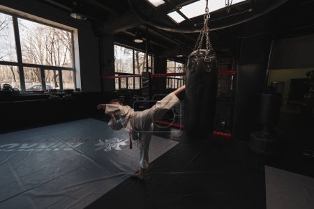 Photo for A girl in a kimono exercises with a punching bag in gym while learning karate martial arts - Royalty Free Image