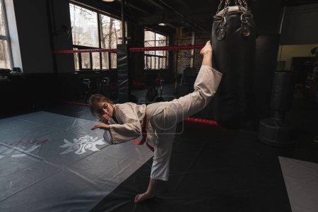 Foto de Una chica en un kimono hace ejercicios con un saco de boxeo en el gimnasio mientras aprende artes marciales de karate - Imagen libre de derechos