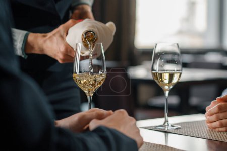 Foto de Una cita en un restaurante de hotel un hombre y una mujer beben vino blanco en copas camarero vierte vino de una botella - Imagen libre de derechos