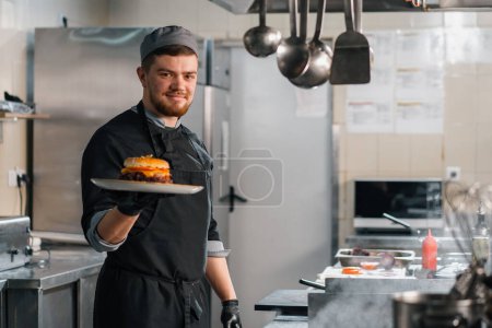 Foto de Cocina profesional en un restaurante del hotel, un chef satisfecho muestra una hamburguesa recién preparada en el plato - Imagen libre de derechos